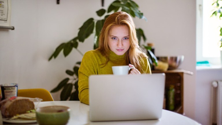 Frau mit rötlichen Haaren und Tasse in der Hand sitzt vor ihrem Laptop am Küchentisch.