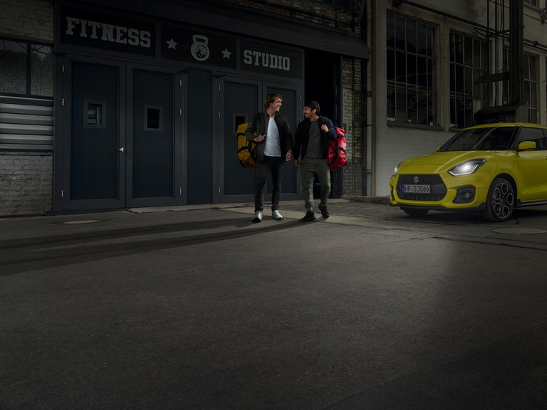 Zwei Männer verlassen ein Fitness Studio, daneben steht ein Suzuki Swift Sport Hybrid in Champion Yellow mit Guide me Light.