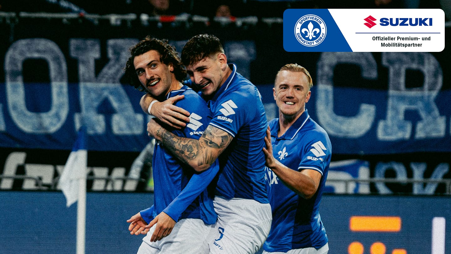 Drei Spieler des SV Darmstadt 98 in blauen Trikots jubeln gemeinsam auf dem Fußballplatz, Sponsor Suzuki im Bild.